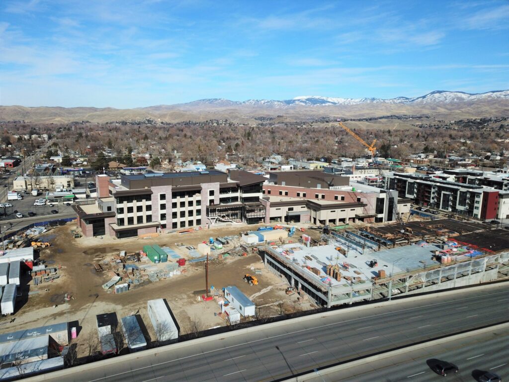 Aerial view of a construction site with cranes near residential area and mountains in the background, under a clear blue sky.