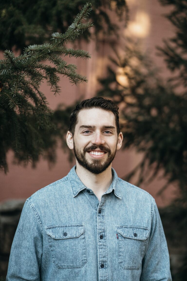 A person with a beard smiles, wearing a denim shirt, standing in front of a background with trees and a brick wall.