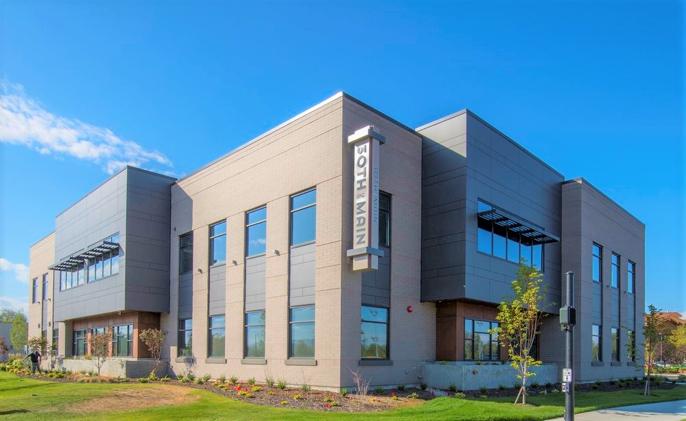 Modern two-story building with large windows and "South Main" sign. Clear blue sky, landscaped greenery, and person walking nearby.