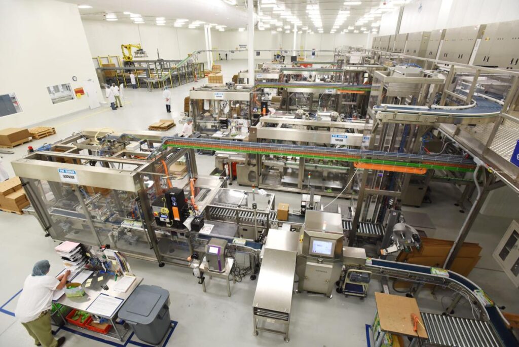 A busy food factory production line with machinery, conveyor belts, and several workers in protective clothing inspecting and managing operations.