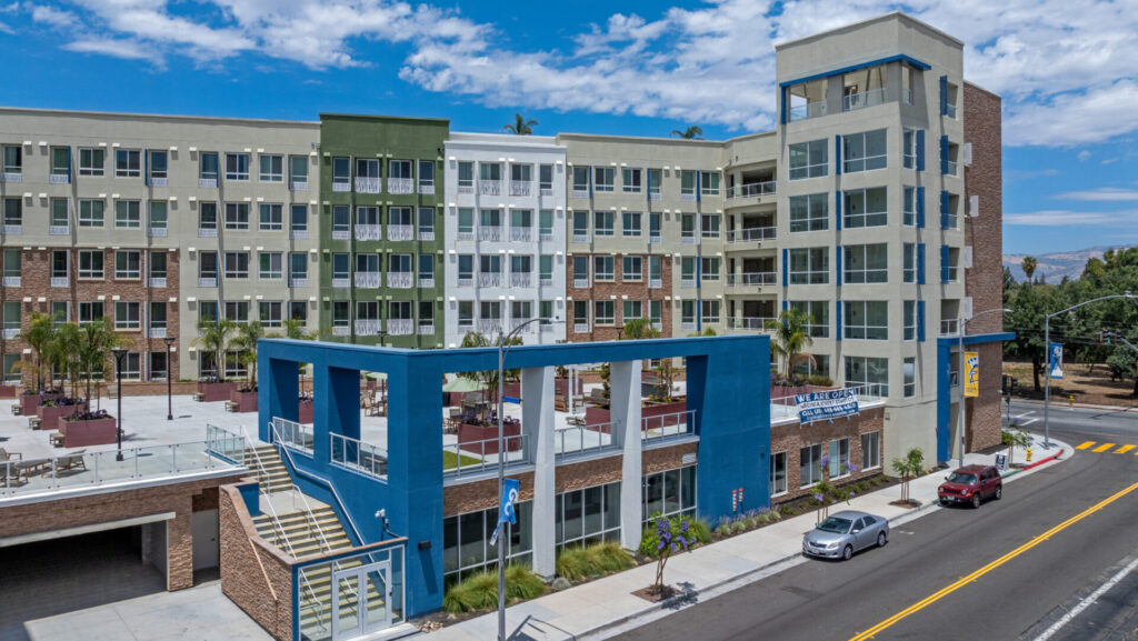 Modern modular multi-story residential apartment building with green, white, and brick facade, featuring an outdoor courtyard with seating, palm trees, and a distinctive blue architectural structure at the entrance along a clean urban street.