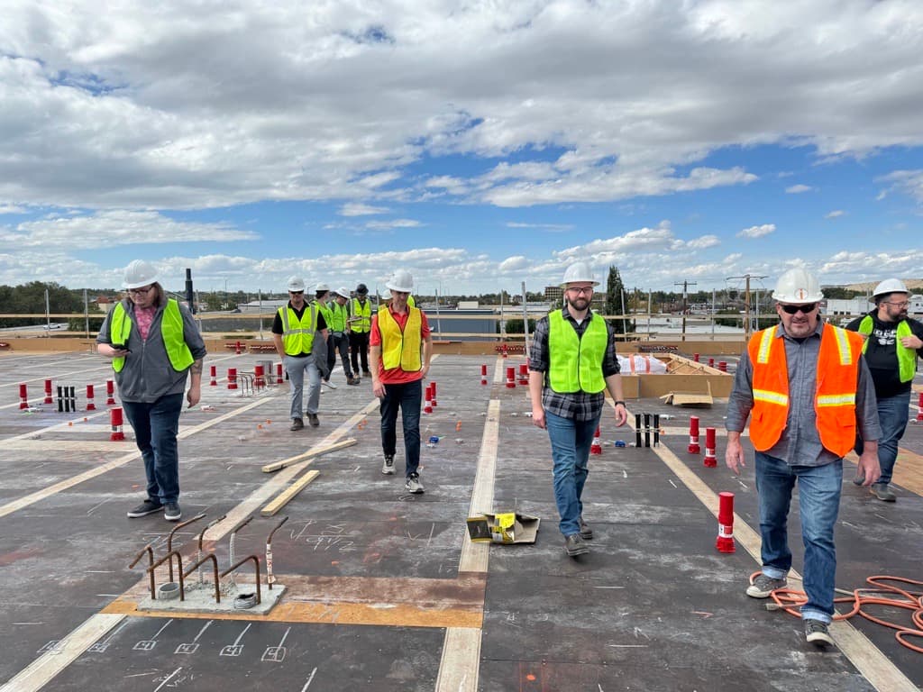 A group of people in hard hats and safety vests walk on a construction site under a partly cloudy sky, surrounded by equipment.