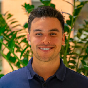 A person smiling in front of green foliage, wearing a navy blue shirt, with a blurred brick background behind them.