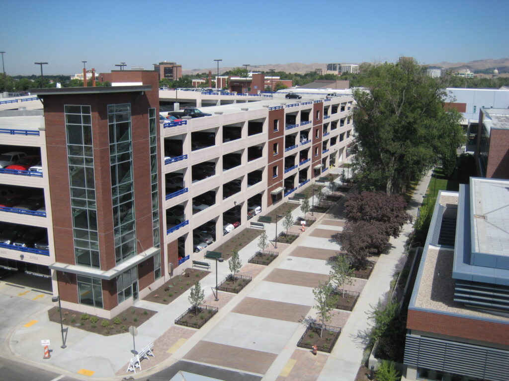 A multi-story parking structure stands beside a landscaped walkway with trees, situated in an urban area under a clear blue sky.