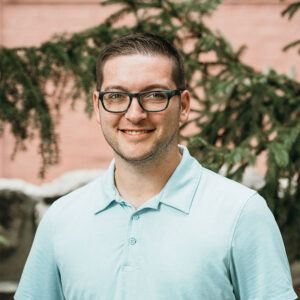 A person with glasses and a light blue shirt smiles in front of a tree with a peach-colored wall background.