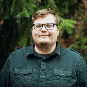 A person with glasses and a green shirt stands in front of lush greenery, smiling slightly, with no visible landmarks or buildings present.