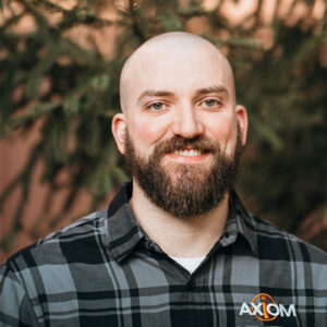 A person with a beard and a plaid shirt smiles outdoors, standing in front of a tree, wearing clothing branded with "Axiom."