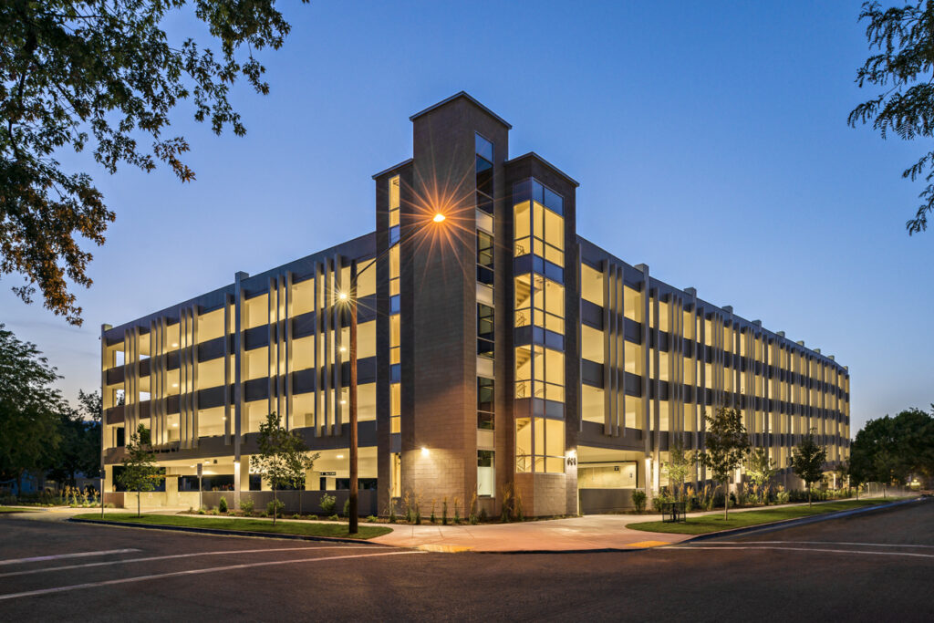 Modern multi-level parking structure illuminated at twilight, surrounded by trees and a quiet street. No people or vehicles are visible.