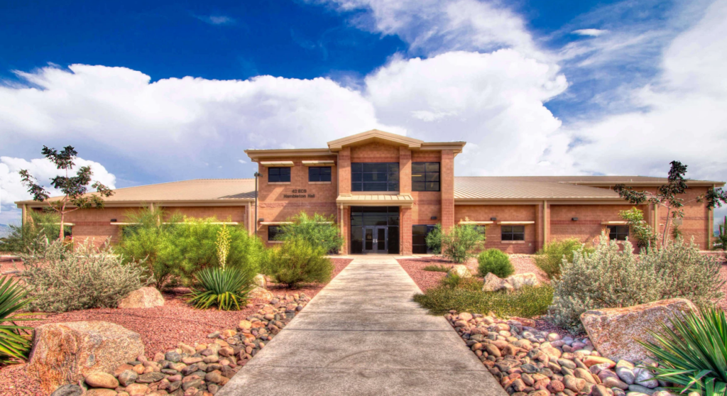 A modern, brick building surrounded by desert landscaping under a partly cloudy sky, with a central entrance pathway leading to the structure.