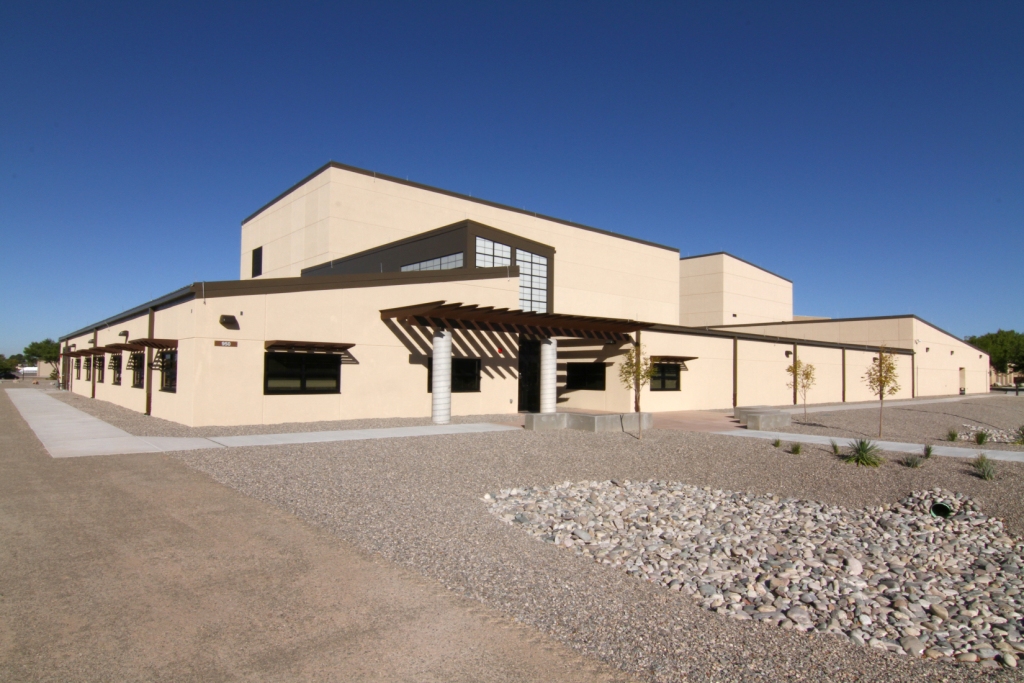 A modern beige building with large windows and a pergola entrance is surrounded by gravel landscaping under a clear, blue sky.