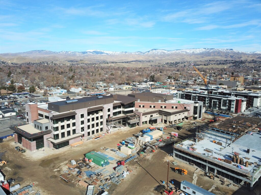 Construction site with several buildings under development, surrounded by a suburban area. Snow-capped mountains are visible in the background under a clear sky.