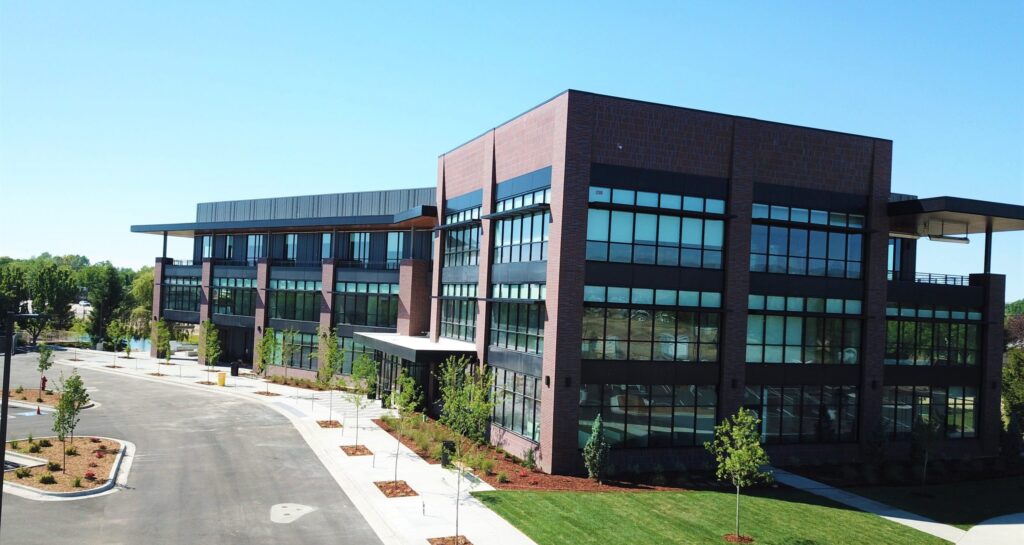 A modern, multi-story office building with large windows, surrounded by neatly landscaped greenery and a spacious parking area under a clear blue sky.