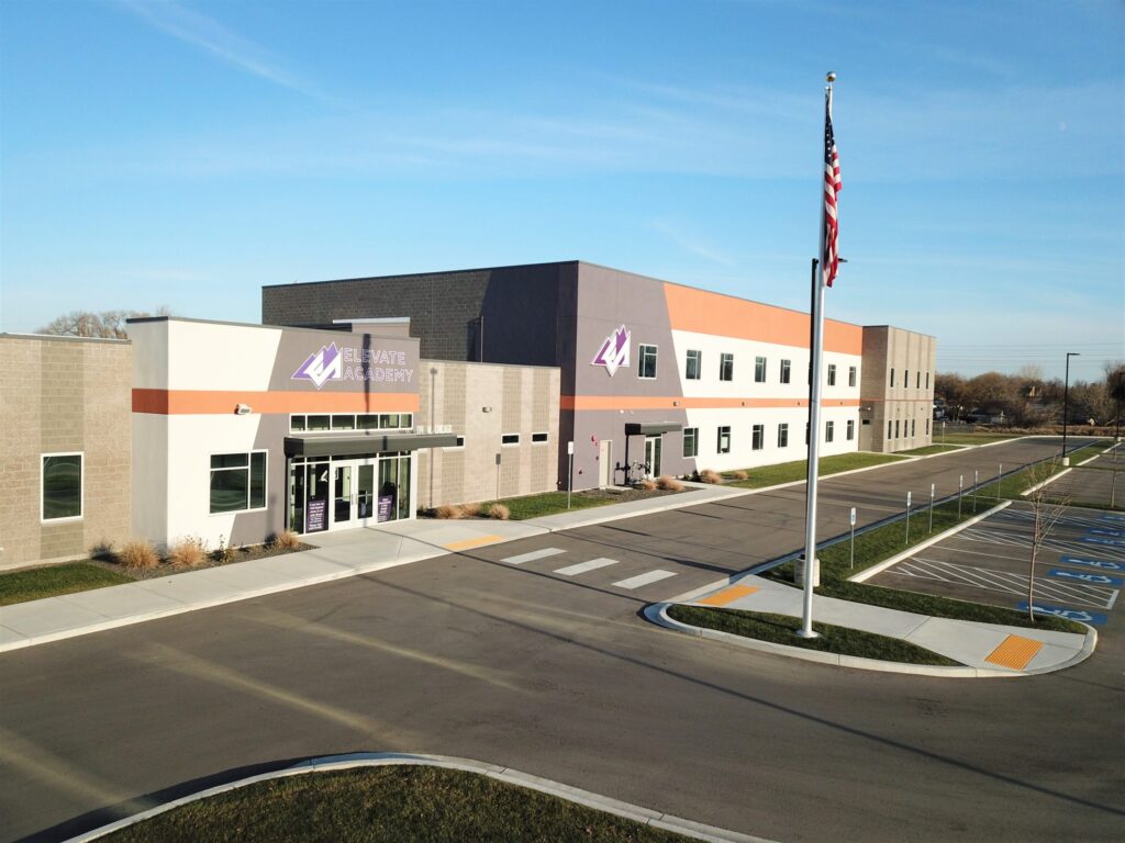 A modern two-story building labeled "Elevate Academy" stands beside an empty parking lot, under a clear blue sky with a flagpole in view.