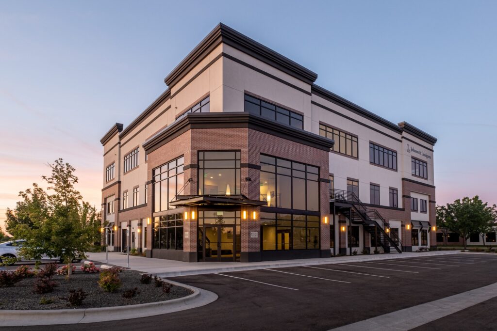 A modern three-story office building with large windows and brick accents, surrounded by a parking lot and trees at sunset.