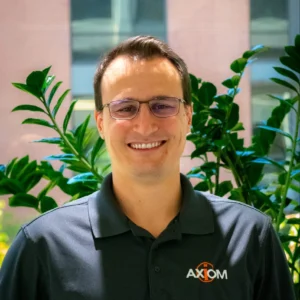 A person with glasses and a polo shirt with the "AXIOM" logo, smiling indoors. Green plants and a building are in the background.