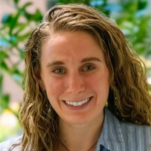 A smiling person with curly hair is in the foreground, set against a blurred background of green foliage.