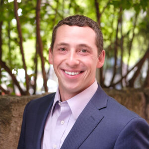 A person in a suit smiles outdoors with a blurred, leafy background. There are no recognizable landmarks or historical buildings.