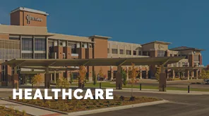 Modern hospital building with clear blue sky. "HEALTHCARE" text overlay. The facility features multiple stories and a spacious entrance with surrounding greenery.