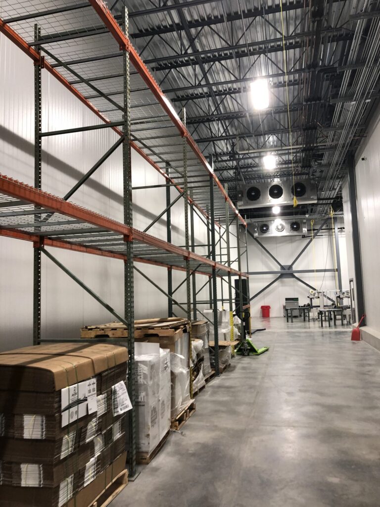 Industrial warehouse interior with metal shelving, pallets, and equipment. High ceiling with lighting and ventilation. Clean, spacious concrete floor. No recognizable landmarks visible.