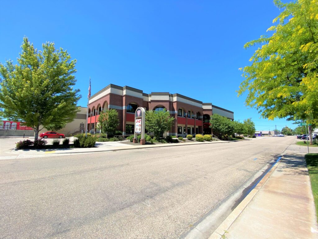 A red brick building with arched windows surrounded by trees and a clear sky. A red car is parked nearby.