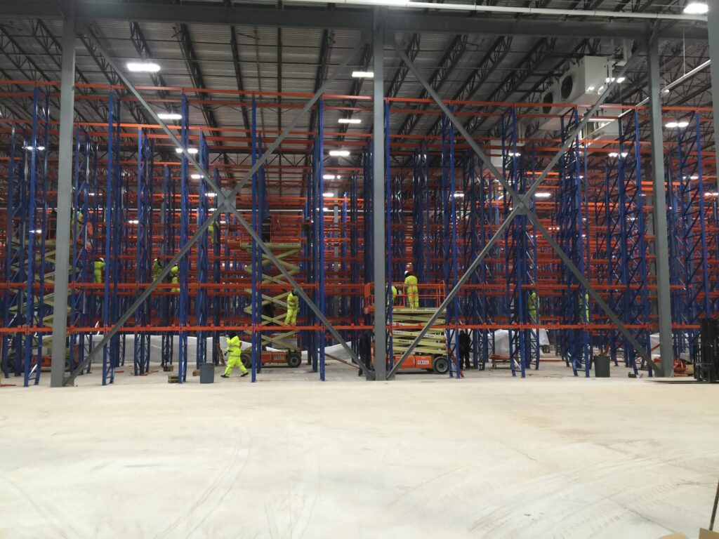 A large warehouse with extensive shelving under construction. Several people in high-visibility clothing are working on scissor lifts, assembling the structure.