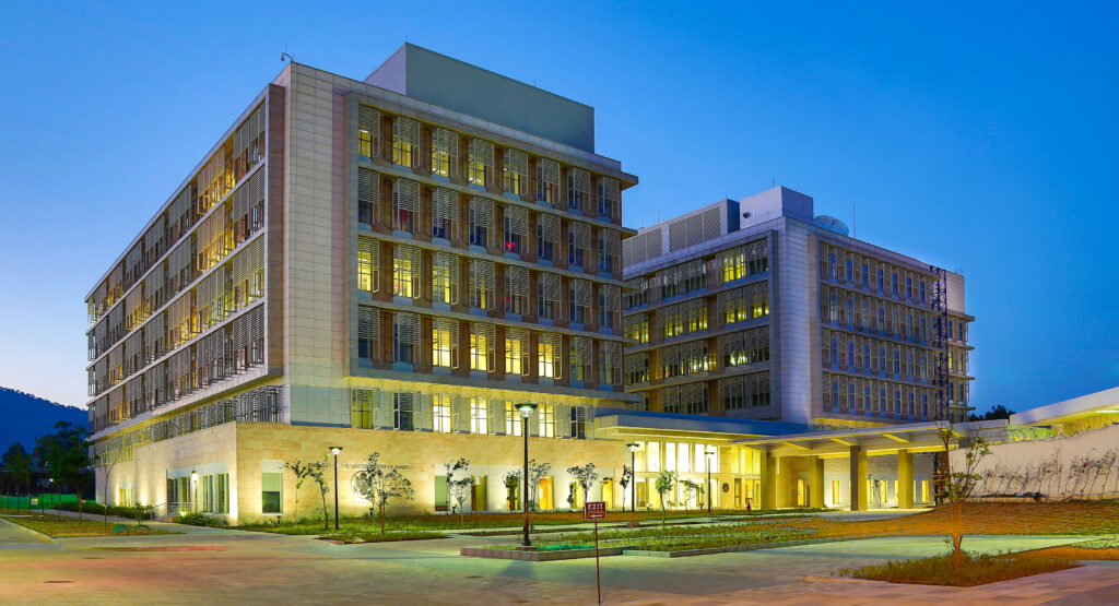 A modern, well-lit office building with multiple floors, surrounded by small trees and lampposts, is depicted against a clear evening sky.