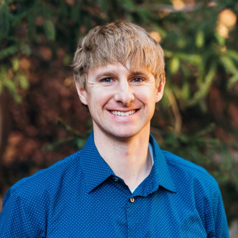 A person with short, light hair smiling and wearing a blue shirt, stands outdoors with blurred greenery in the background.