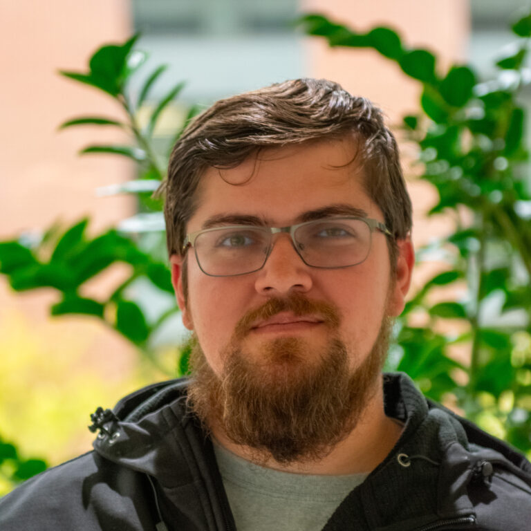 A person with glasses and beard is in focus, with green leaves and a brick wall blurred in the background.