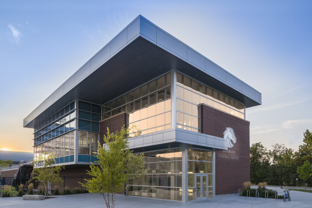 Modern building with large glass windows, brick walls, and metal accents. Located outdoors with trees and clear sky in the background.