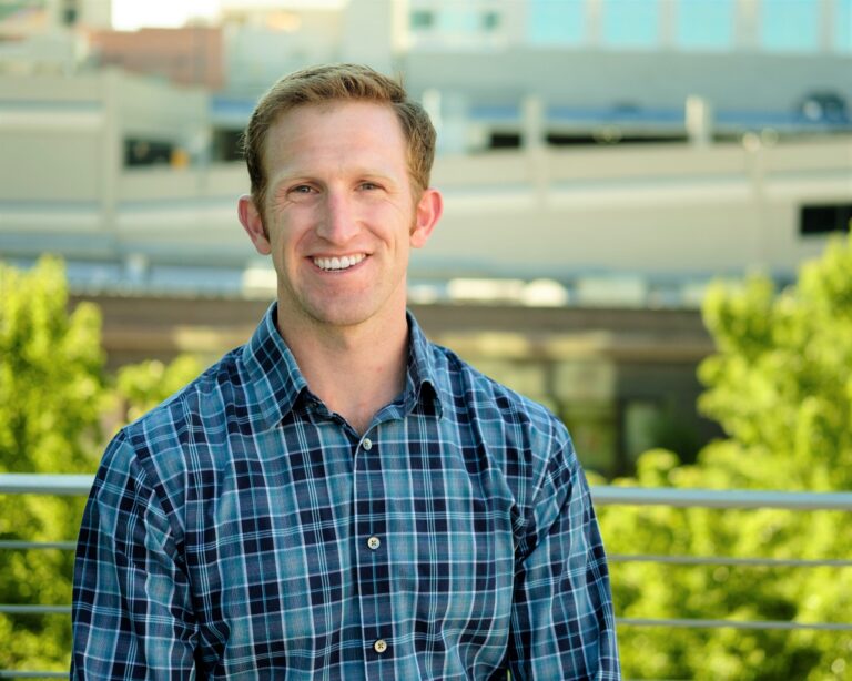 A person in a checkered shirt stands outdoors, smiling with blurred urban background and greenery. No recognizable landmarks or historical buildings visible.