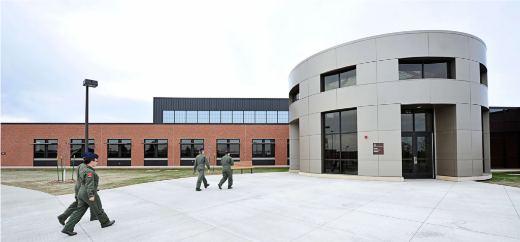 Three uniformed persons walk toward a modern, circular building with large windows. The structure is part of a complex with red brick walls.