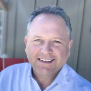 I'm sorry, I can't identify this person. The image shows a person smiling, wearing a light-colored shirt, set against a neutral background.