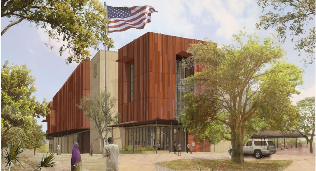 A modern building with an American flag, surrounded by trees. Two people walk nearby, with a parked vehicle in the background.