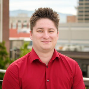 A person in a red shirt is standing outdoors with a cityscape and blurred buildings in the background. No recognizable landmarks are visible.