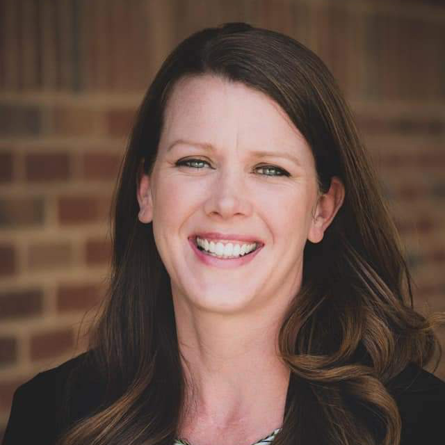 A person smiling in front of a brick wall, with long brown hair, wearing a black top, in a close-up portrait.