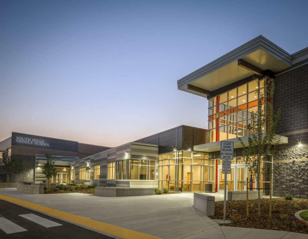 Modern school building exterior at dusk, labeled "South Hills Middle School," with well-lit windows, contemporary architecture, and landscaped entrance without visible people.