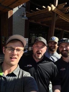 Four people in casual attire take a selfie outdoors. Wooden structures are visible in the background. No recognizable landmarks or historical buildings are present.