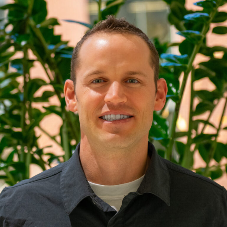 A person with short hair smiles in front of lush green plants, wearing a dark shirt. The background is softly blurred.