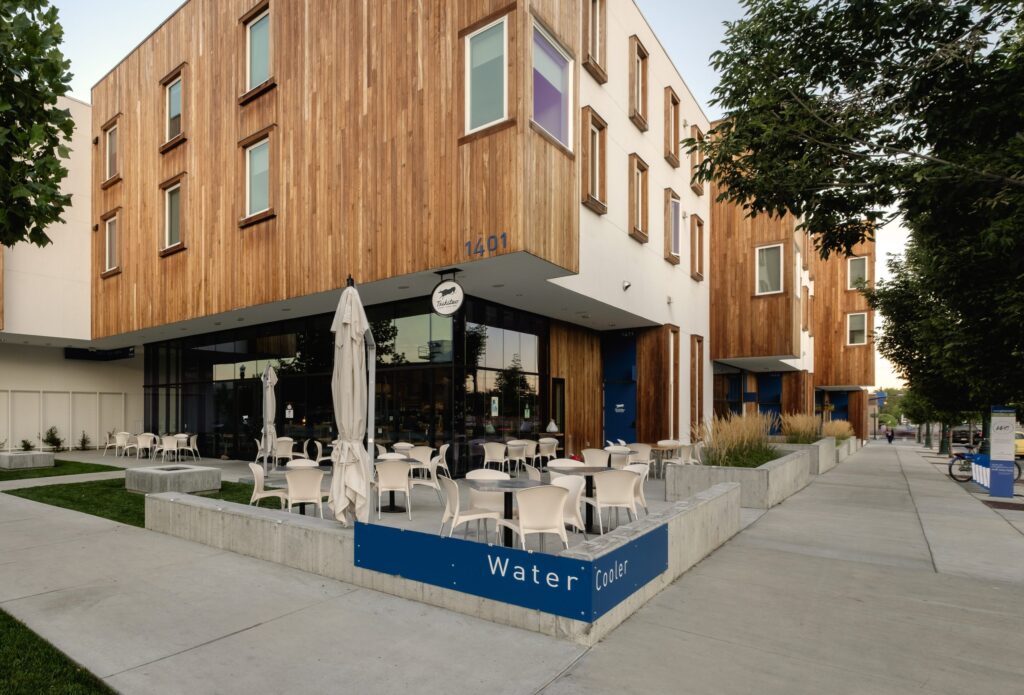 Modern building with wooden facade, outdoor seating area, umbrellas closed. Sidewalk and greenery along the street. No recognizable landmarks visible.