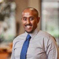 A person wearing a gray shirt and blue tie is smiling indoors, with a blurred background that suggests a professional setting.