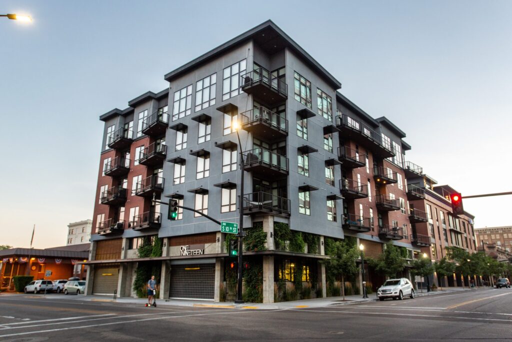 A modern multi-story apartment building on a street corner, with balconies and trees below. A person walks nearby, and cars are parked.