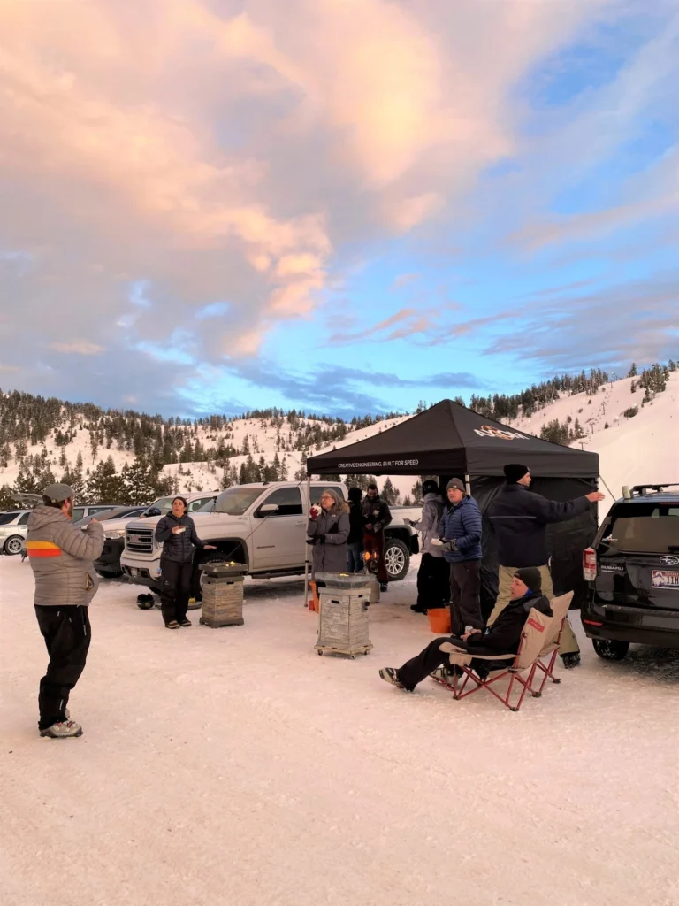 A group of people gathers near trucks and a black tent in a snowy mountainous area under a colorful evening sky.