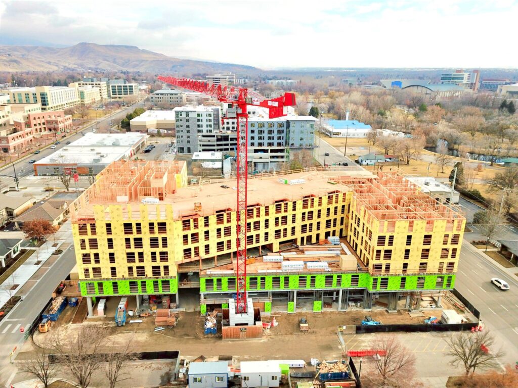 A large building under construction with a red crane, surrounded by roads and urban scenery, set against a backdrop of mountains and trees.