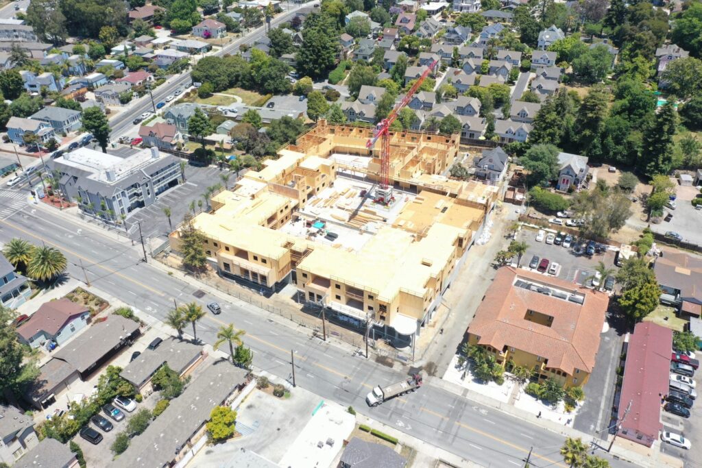 Aerial view of a construction site with a crane in a suburban neighborhood. Surrounding streets, trees, and houses are visible.