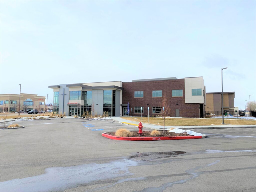 Modern, two-story building with large windows in a mostly empty parking lot. Sky is clear, with a fire hydrant and minimal landscaping visible.