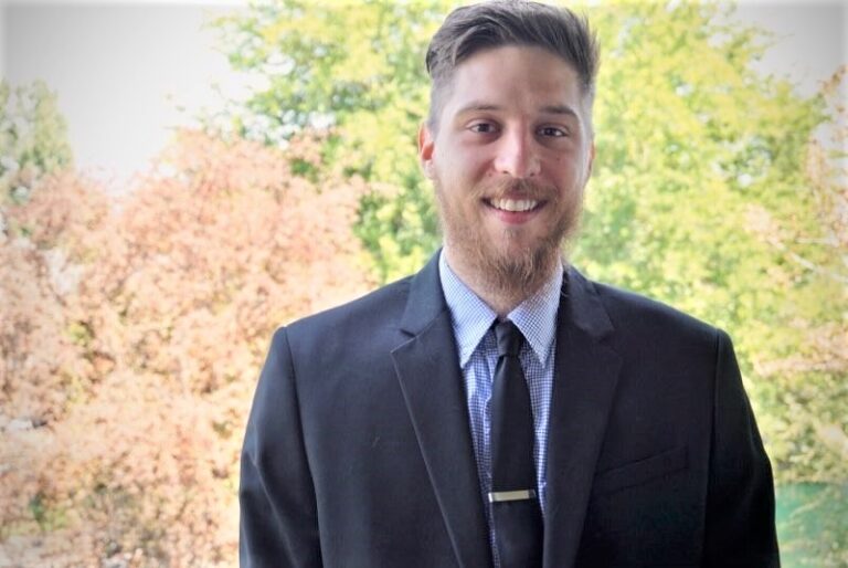 A person in a suit smiles in front of lush greenery and blurred trees, conveying a formal yet natural setting.