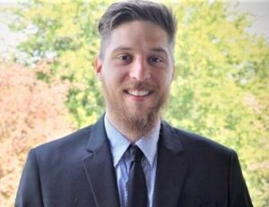 A person in a suit smiles in front of lush greenery and blurred trees, conveying a formal yet natural setting.