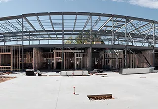 A building under construction with a metal framework, set against a clear sky. Construction materials and equipment are scattered on the site.