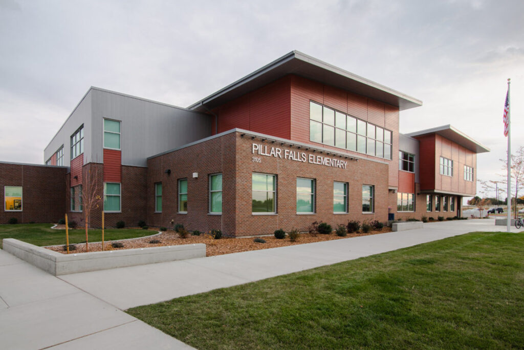 Modern brick building labeled "Pillar Falls Elementary," with large windows, manicured landscaping, and an American flag on a flagpole in front.