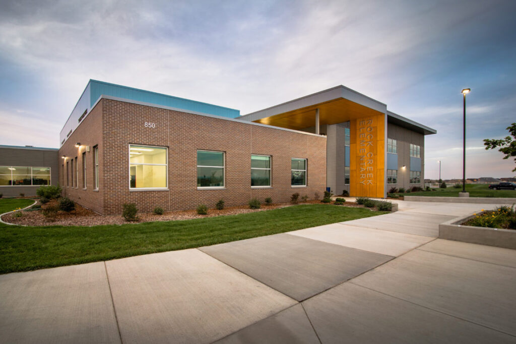 Modern school building with brick façade, large windows, and "Rock Creek Elementary" signage. Surrounded by landscaped lawns, under a clear evening sky.